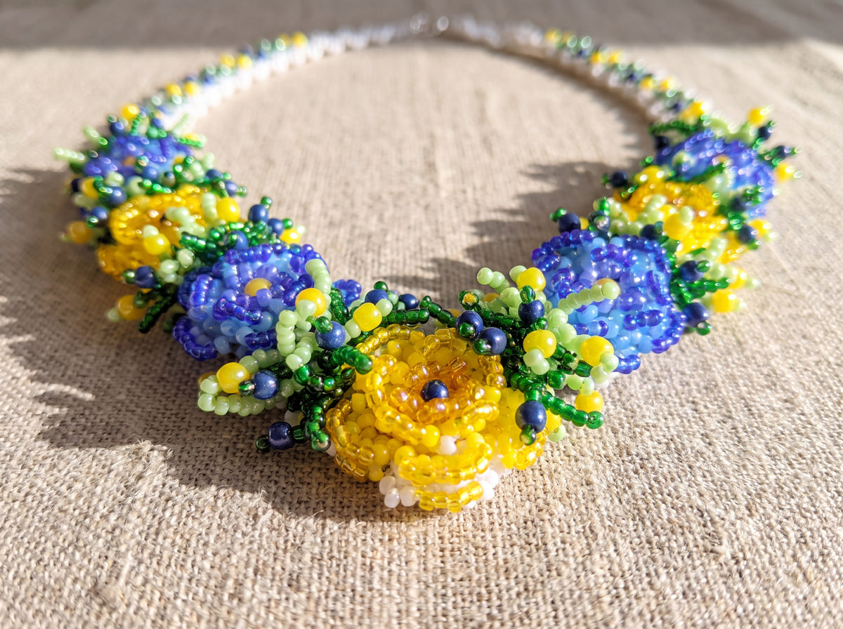 Close-up of a handmade beaded necklace with dimensional blue and yellow seed-bead flowers and green sprigs, laid flat on natural linen in sunlight