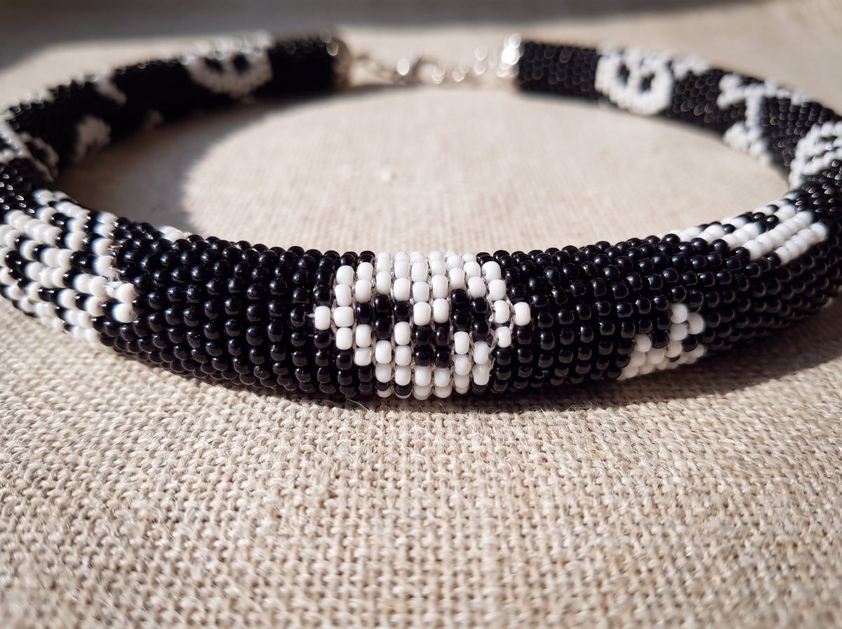 Close-up detail of a handmade black and white seed-bead crochet necklace with skull pattern, resting on natural linen