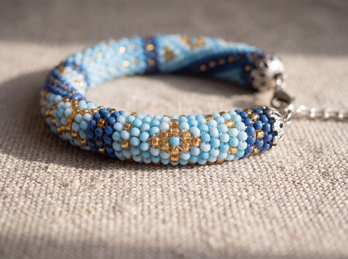 Close-up detail of a handmade bead-crochet bracelet in light blue, navy and gold seed beads with silver clasp, on natural linen