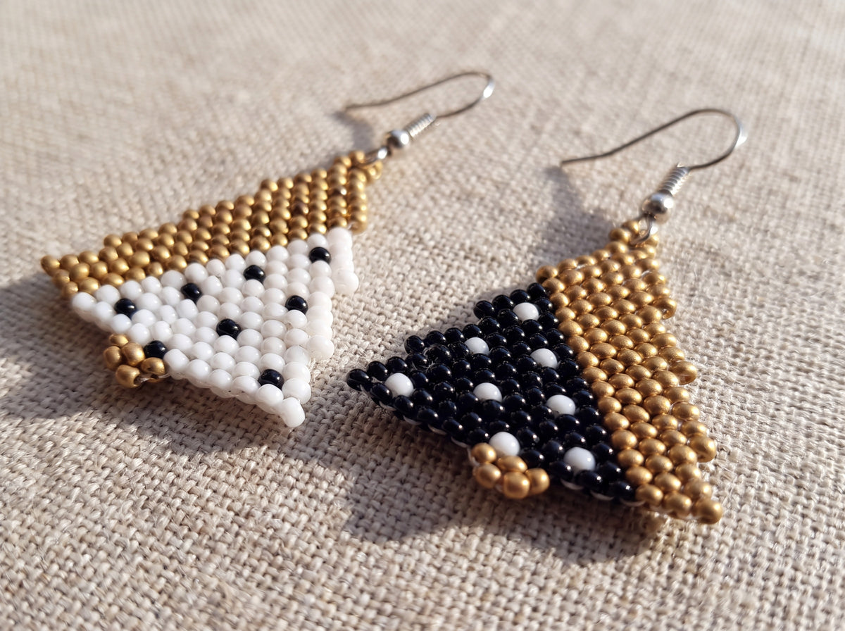 Close-up of handmade beaded Christmas tree earrings, one white and one black with polka dots and matte gold accents, laid on linen