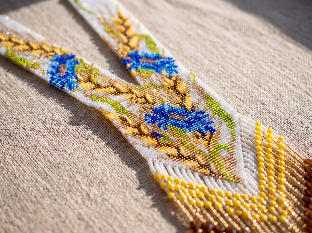 Close-up detail of a handmade beaded gerdan showing wheat and blue cornflower pattern with yellow and amber fringe on linen fabric.