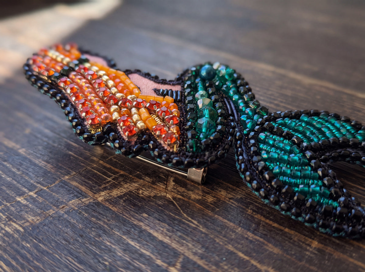 Close-up detail of a handmade beaded mermaid brooch showing orange crystal hair and shimmering teal tail beadwork on dark wood.