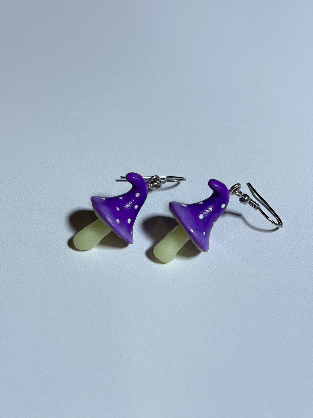 Handmade polymer clay fairy mushroom earrings with purple white-dotted caps on silver hooks, flat lay