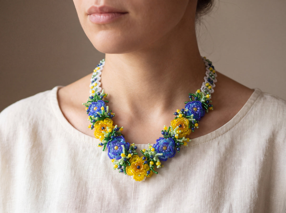 Handmade beaded 3D flower necklace with blue and yellow blossoms and green leaves, worn on a model over a cream linen top