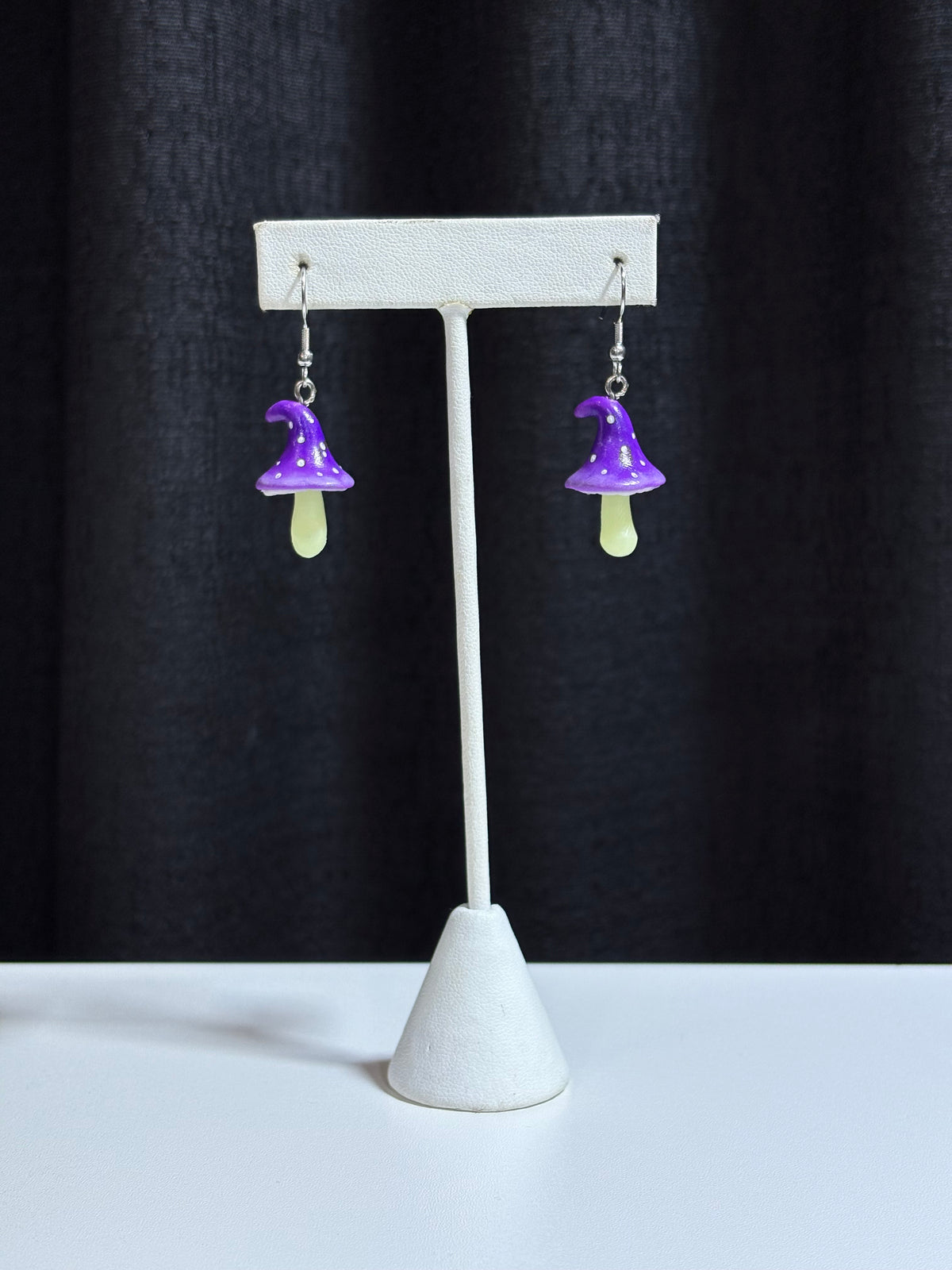 Fairy Mushrooms earrings with purple polka dot caps hanging on a white earring stand against black backdrop
