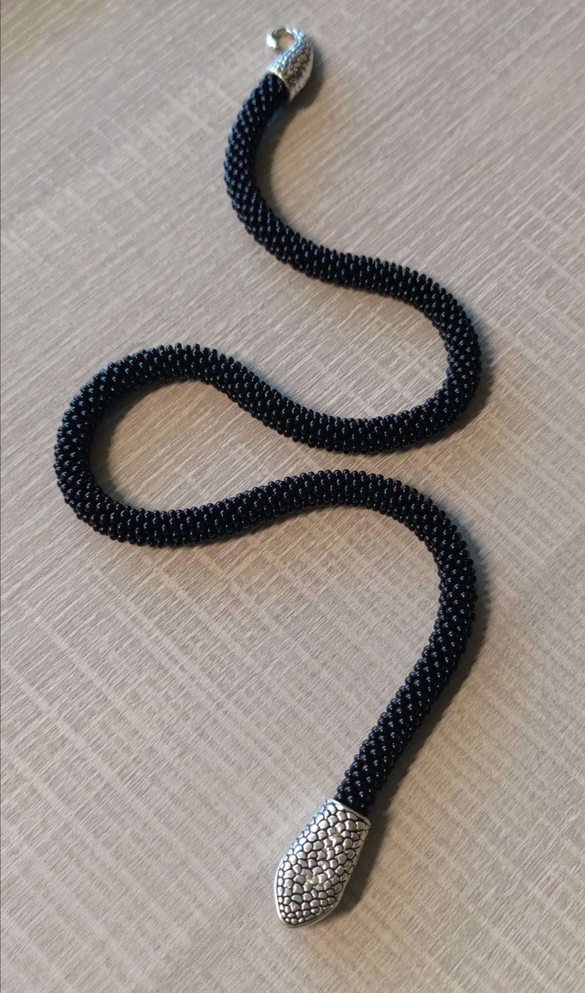 Handmade black beaded snake choker in an S-curve on gray linen, detailed silver snake head clasp