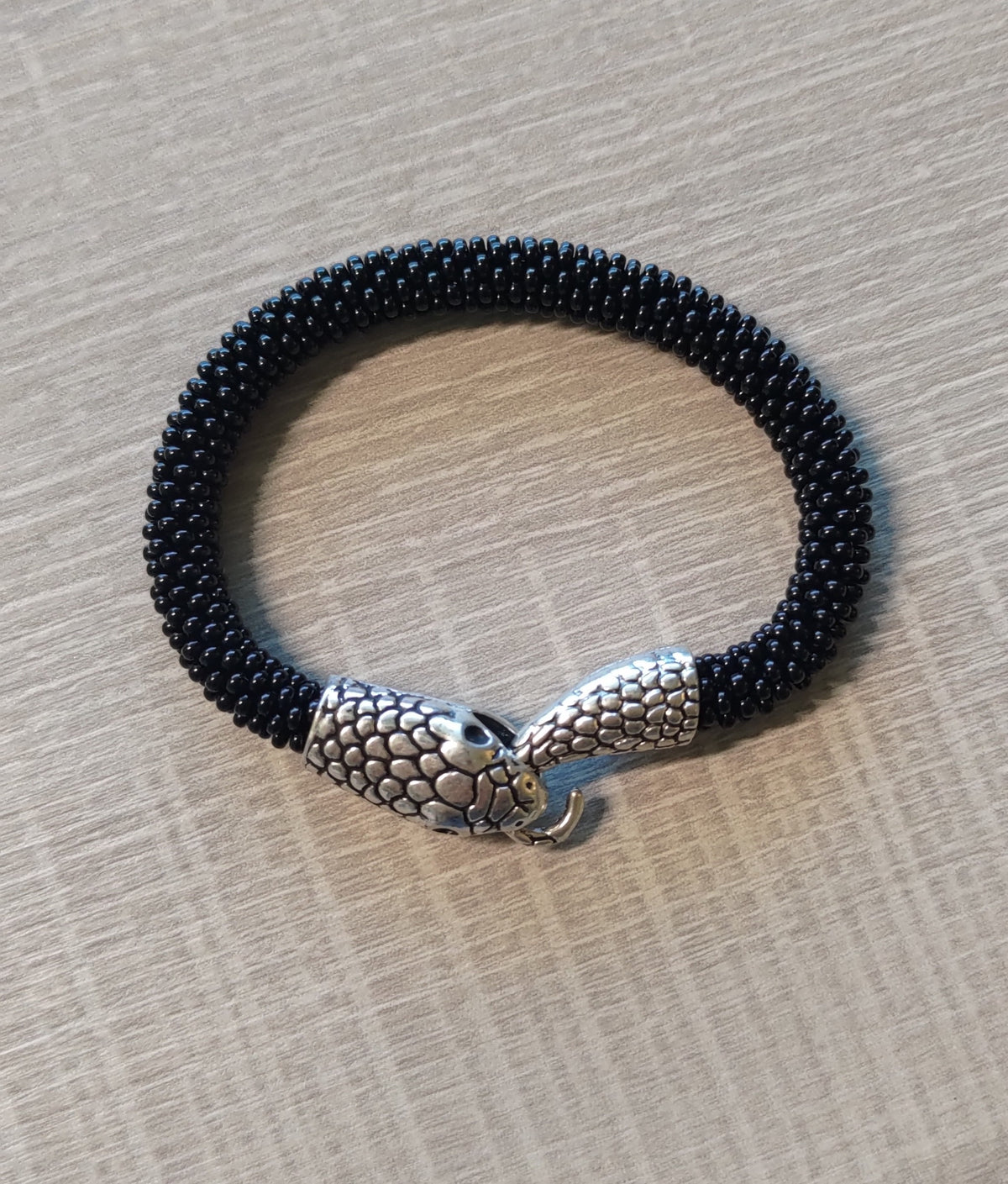 Black beaded snake bracelet crocheted from glass seed beads, silver scaled snake head clasp, on gray tabletop