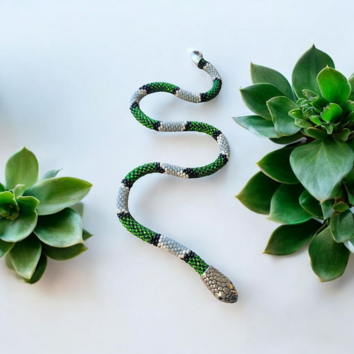 Green, gray and black beaded snake necklace coiled flat between succulents, silver scaled head detail