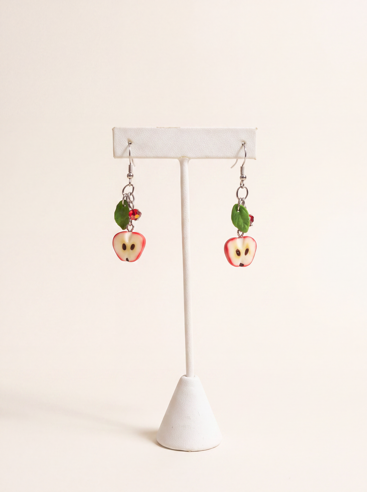 Polymer clay apple-half earrings with green leaves and red crystals on silver hooks, on a display stand