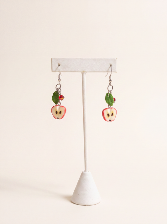 Polymer clay apple-half earrings with green leaves and red crystals on silver hooks, on a display stand