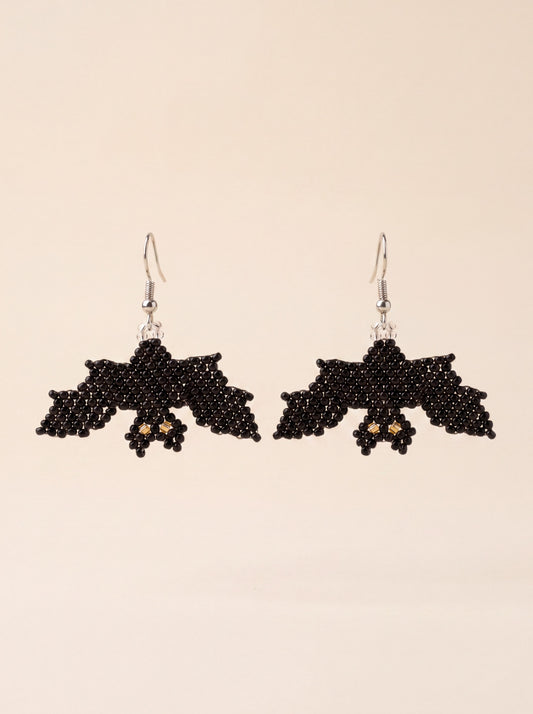 Handmade black beaded bat earrings with gold eyes hanging side by side on silver hooks, on cream