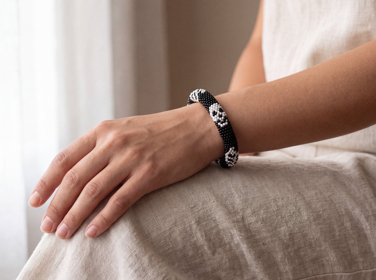 Handmade black bead-crochet bracelet with white skull motifs worn on a model's wrist against cream linen