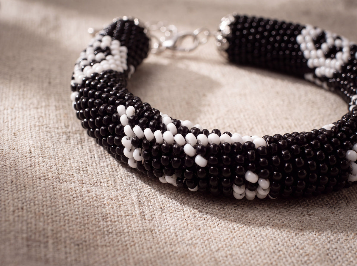 Close-up of a handmade black seed-bead rope bracelet with white skull pattern and silver clasp, laid on natural linen fabric