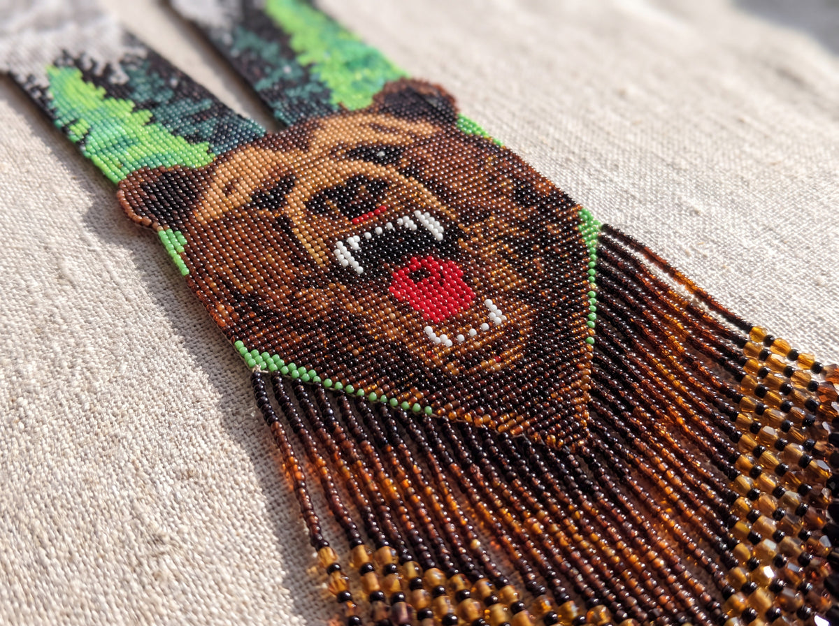 Close-up detail of a beadwoven gerdan necklace showing a roaring brown bear motif and long amber bead fringe on natural linen.