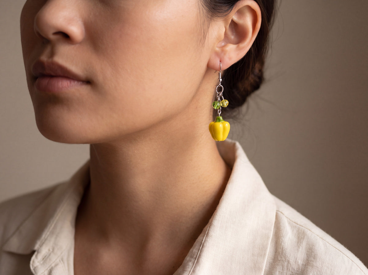 Handmade yellow bell pepper dangle earring with green crystal beads on a silver hook, worn on a model's ear.