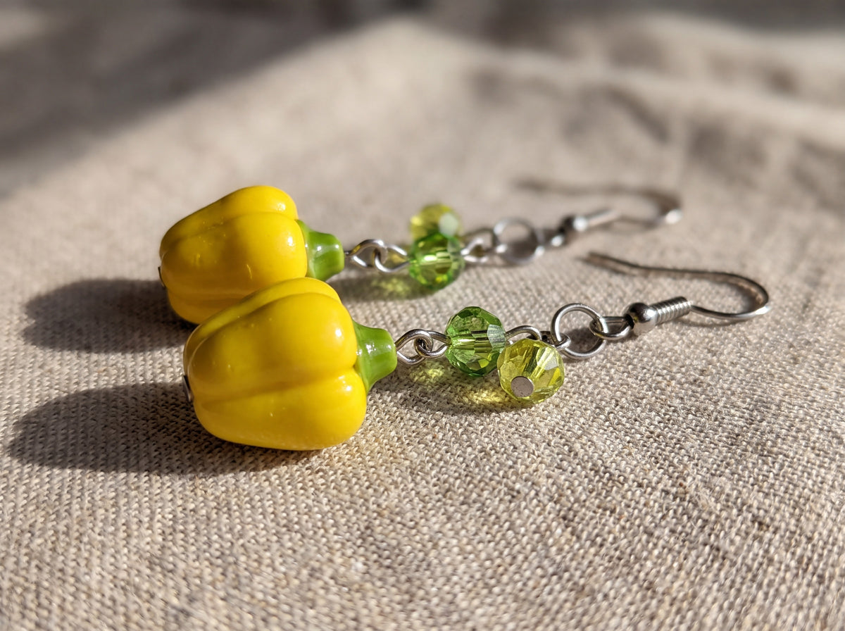 Close-up of handmade yellow bell pepper earrings with green faceted crystal beads and silver hooks resting on natural linen.
