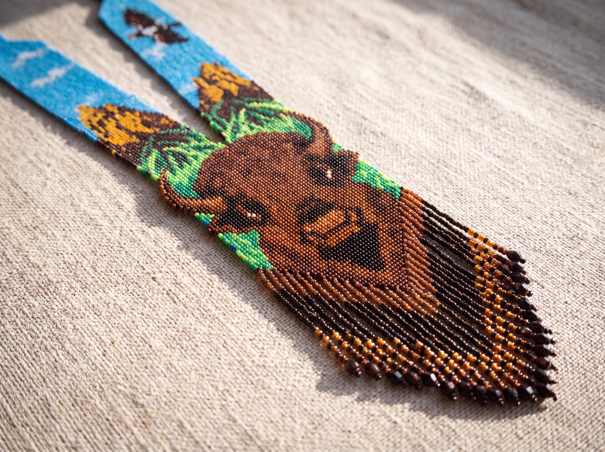 Close-up of a handmade loom-beaded gerdan necklace with buffalo portrait, blue sky and amber fringe, laid on natural linen in sunlight