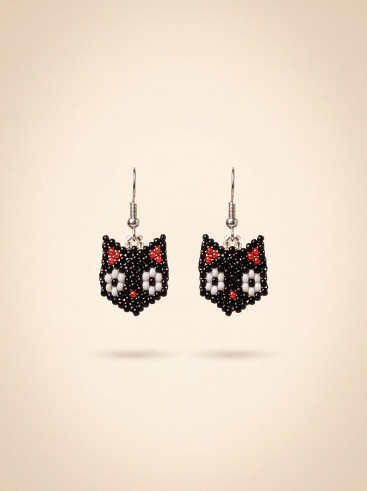 Small handmade black beaded cat earrings with white flower details hanging side by side on silver hooks, on cream