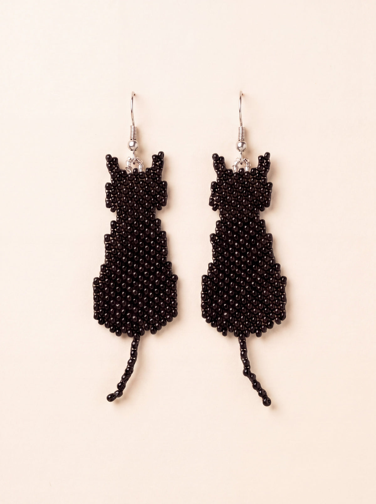 Handmade black beaded sitting cat silhouette earrings with beaded tails hanging side by side on silver hooks, on cream