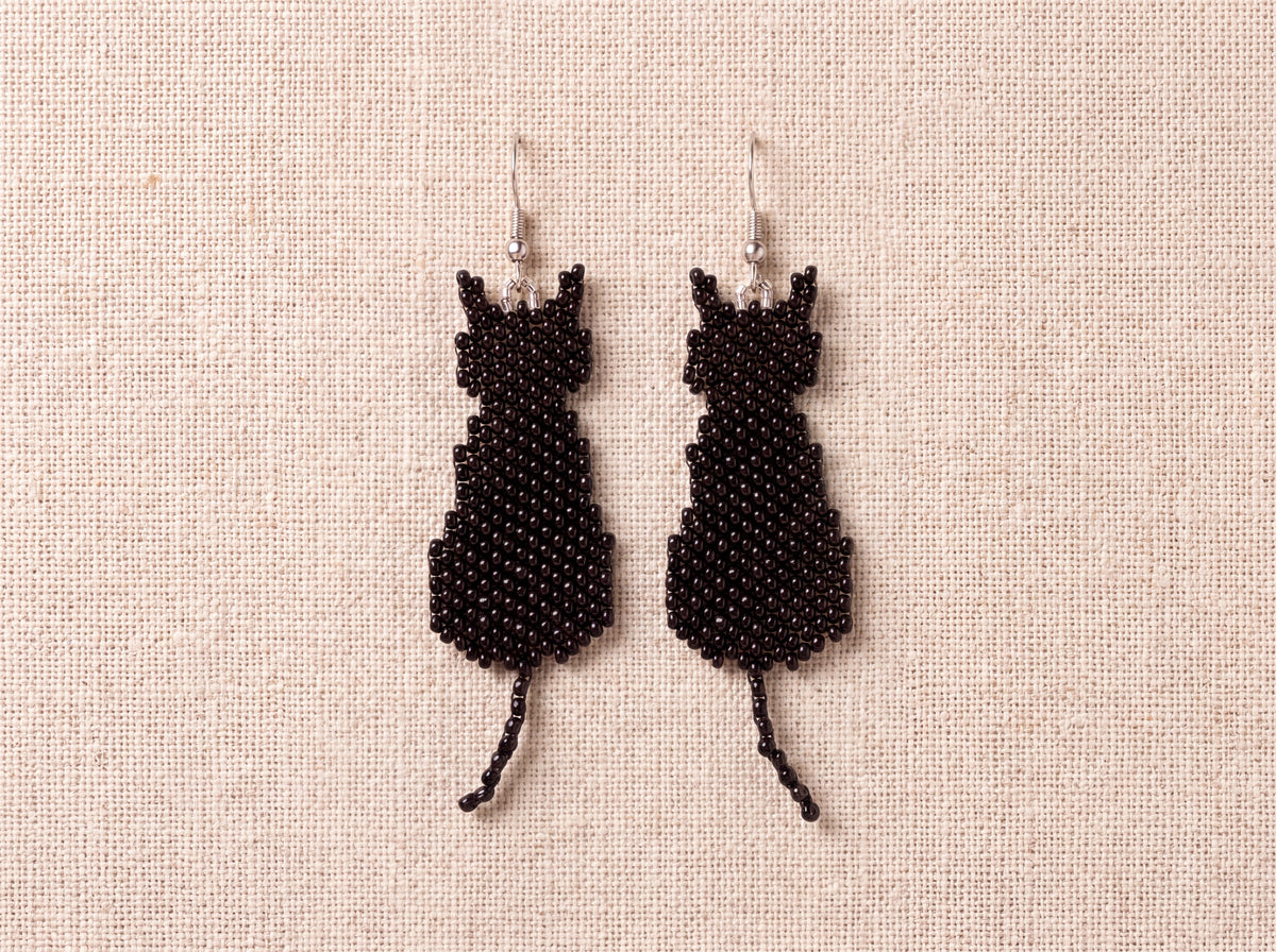 Pair of black beaded sitting cat earrings laid side by side on oatmeal linen