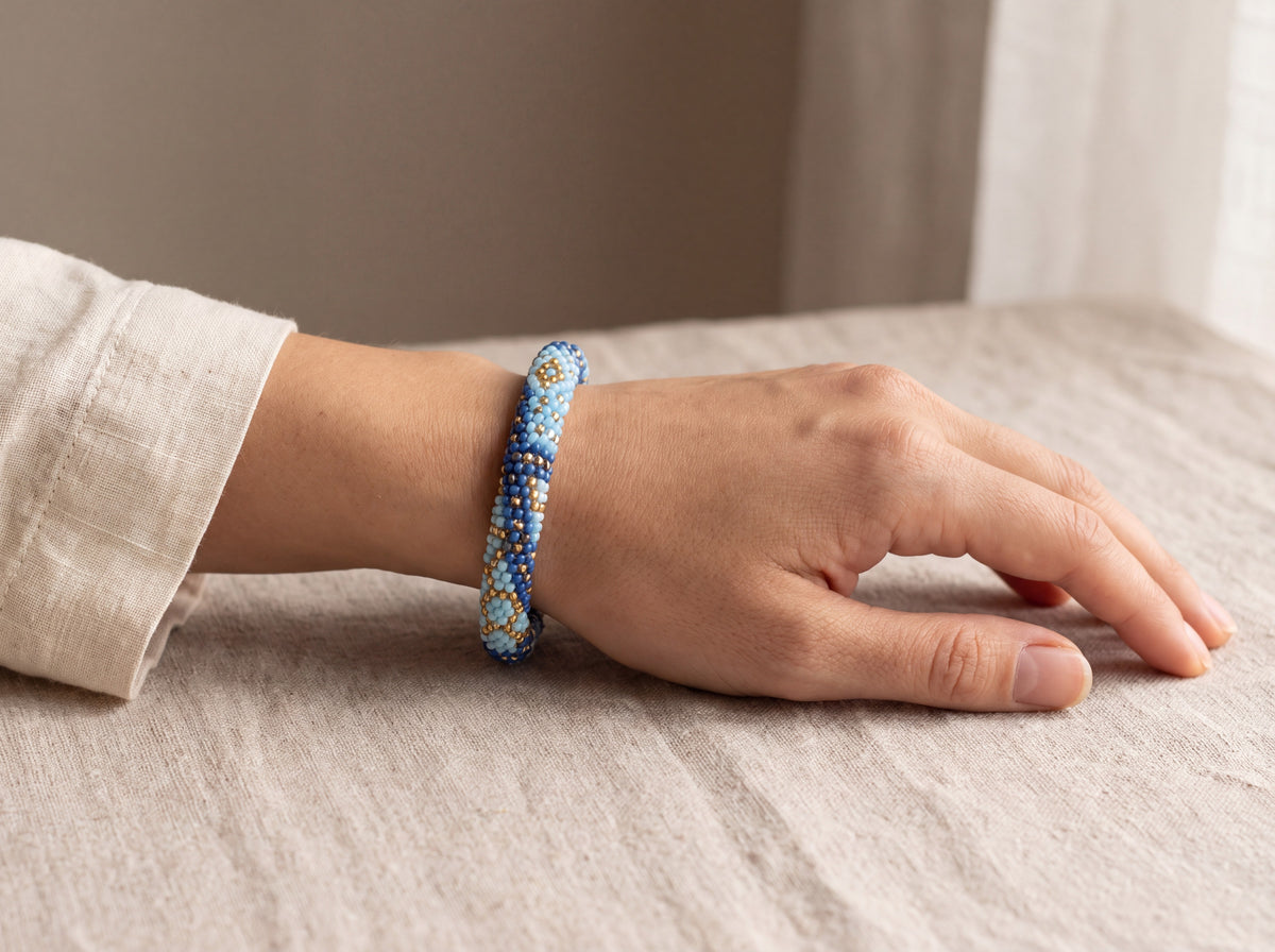 Handmade bead-crochet rope bracelet in navy, light blue and gold worn on a model's wrist resting on beige linen