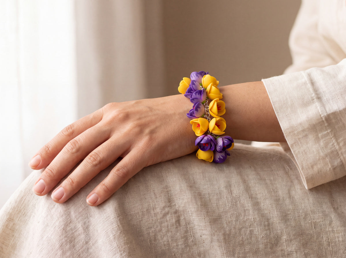 Handmade charm bracelet with purple crocus and yellow flower buds on a silver chain, worn on a woman's wrist against cream linen.
