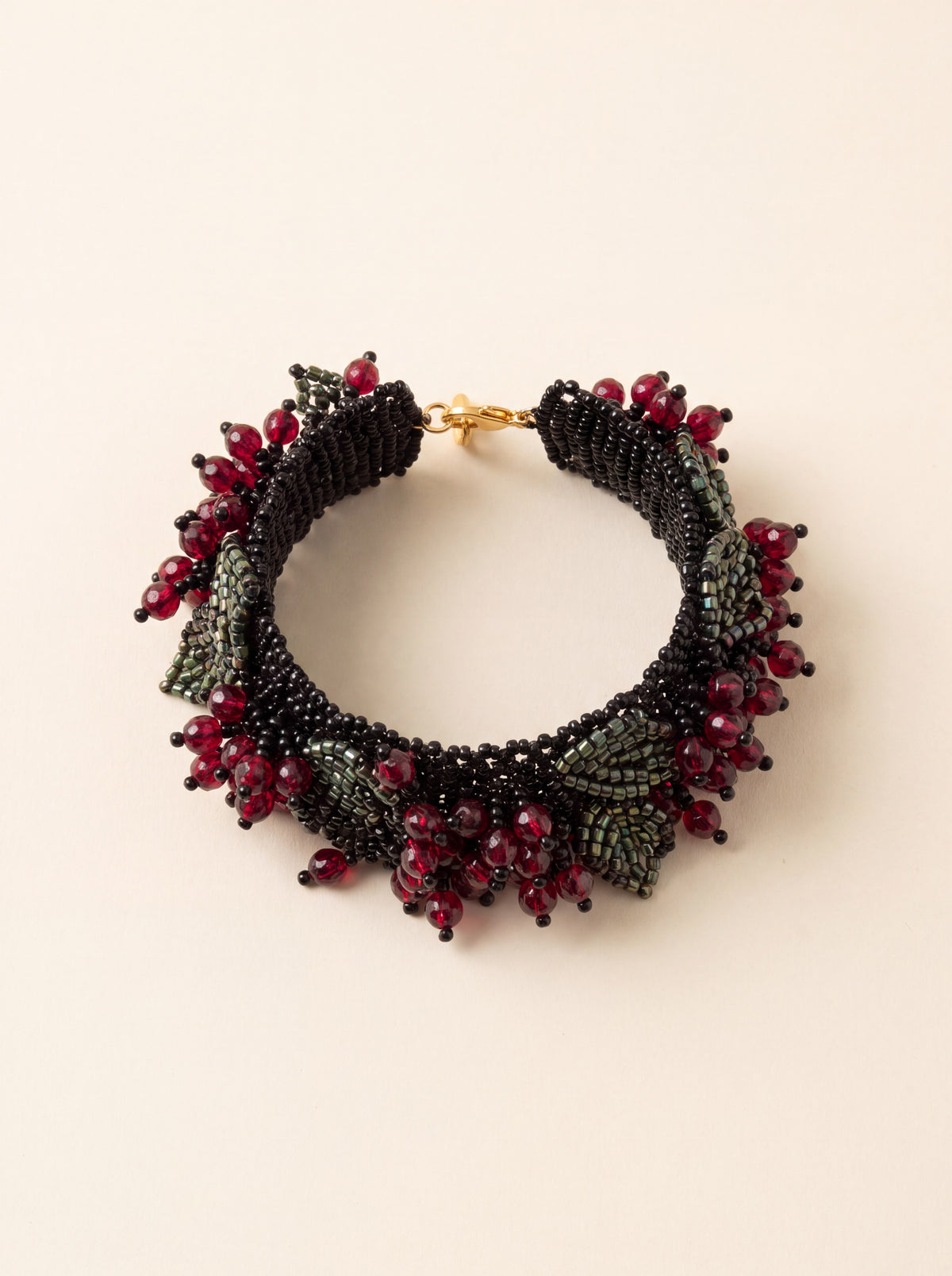 Handmade beaded cuff bracelet with black beadwork lace, garnet-red berries and green beaded leaves, coiled flat on cream