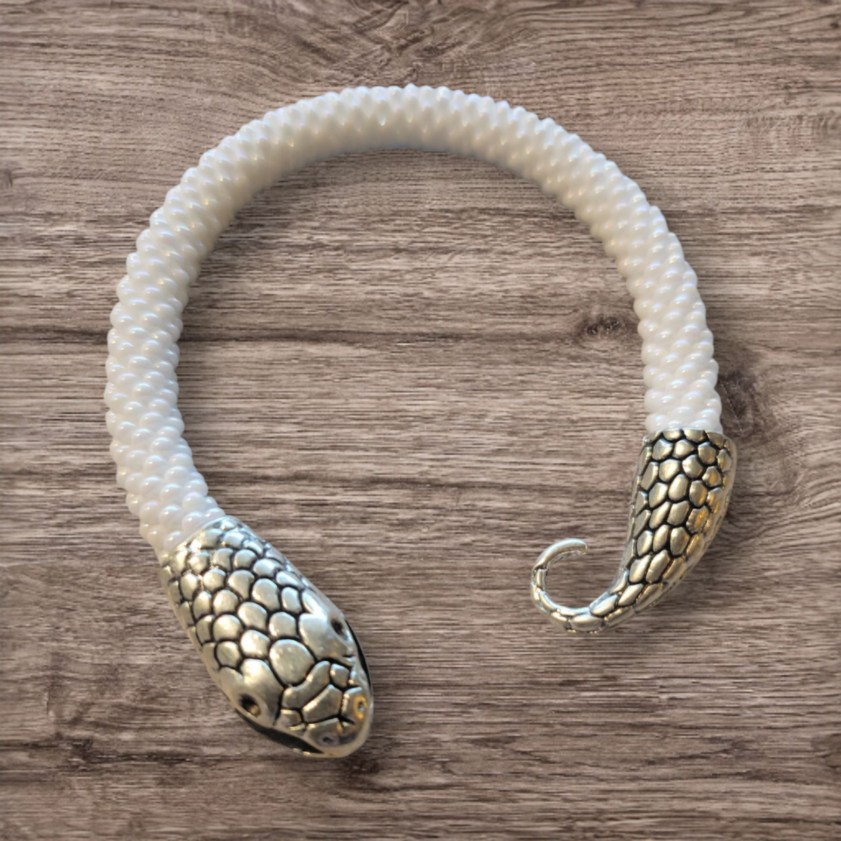 White bead crochet snake bracelet with silver snake head and tail clasp, shown as an open cuff on wood grain