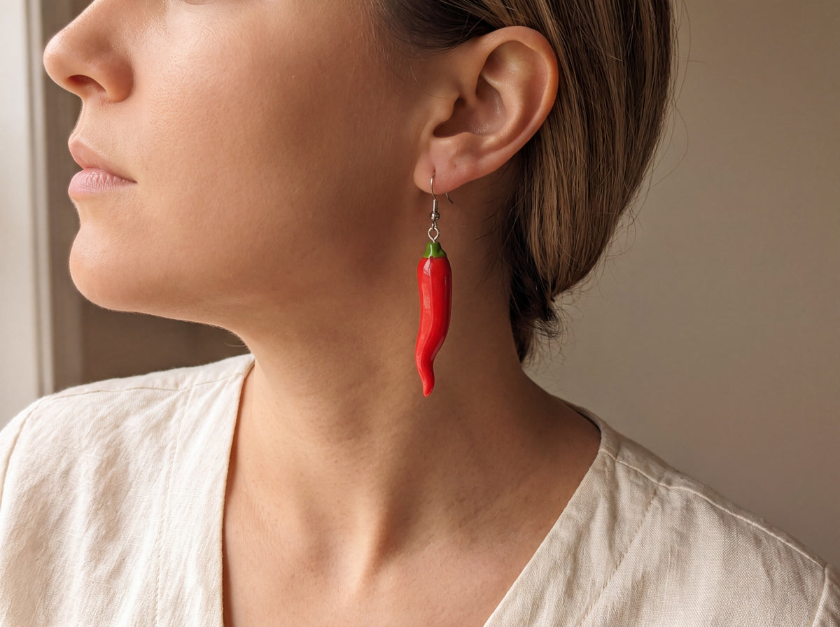 Handmade red chili pepper dangle earring with a green stem on a silver hook, worn on a model's ear in profile.