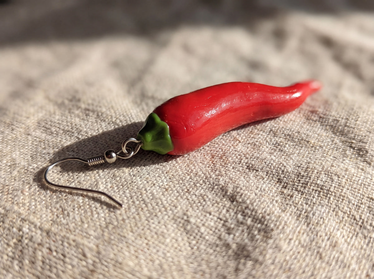 Close-up of a handmade glossy red chili pepper earring with green stem and silver hook lying on natural linen fabric.