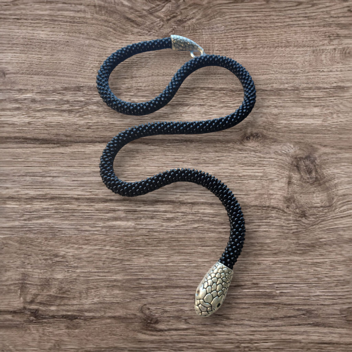 Black glass seed bead snake necklace curving across a wooden background, silver snake head at the tip