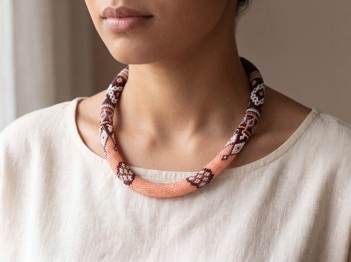 Handmade bead crochet rope necklace in peach, brown and white patchwork patterns, worn around the neck of a model in a cream top.