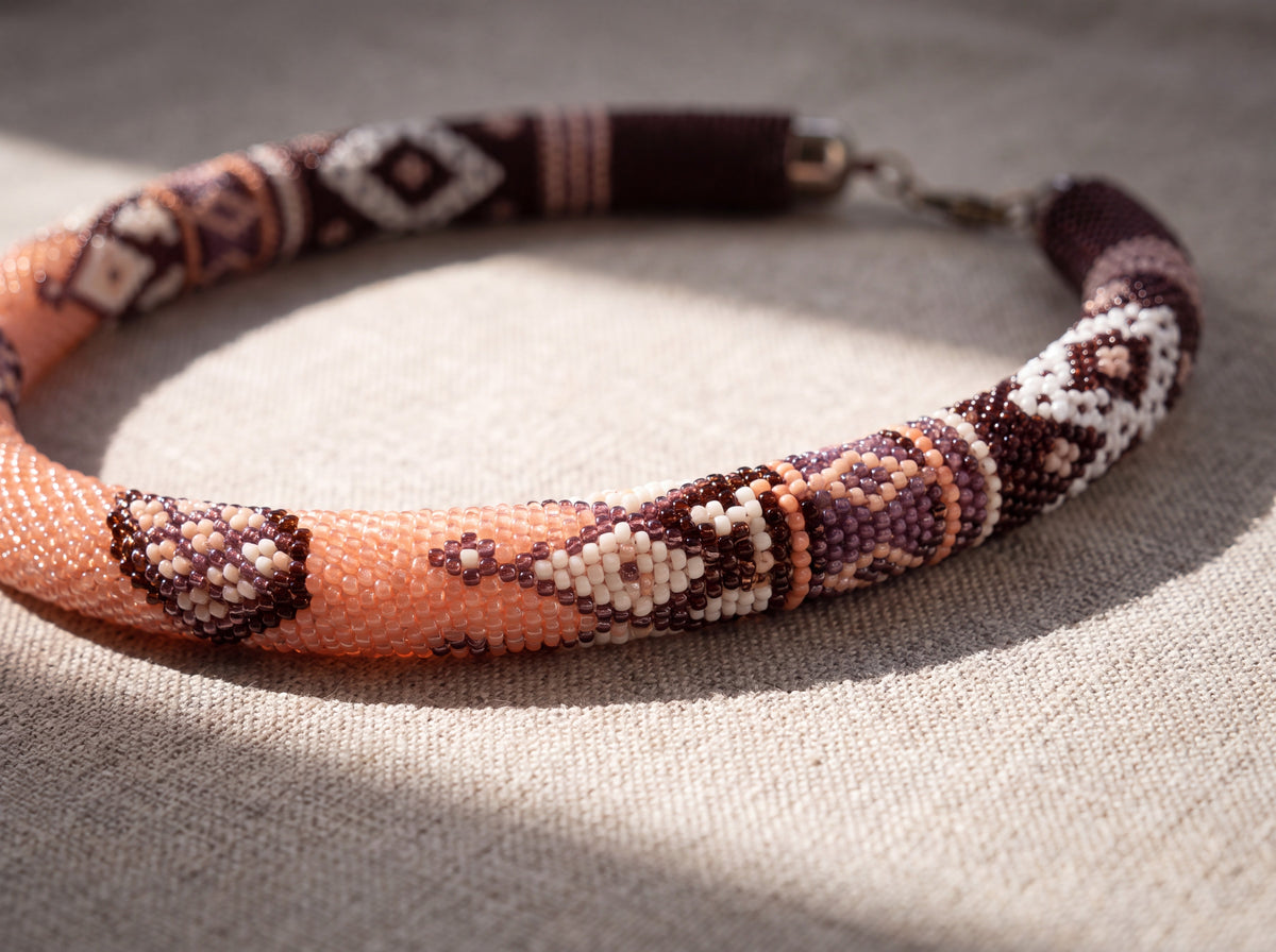 Close-up of a handmade bead crochet coffee-toned rope necklace with peach, cream and brown geometric sections and silver clasp on linen.