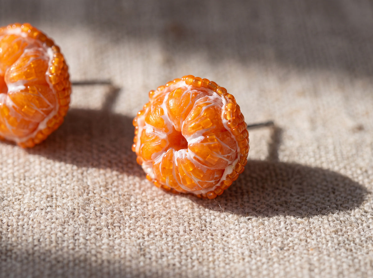 Macro close-up of handmade peeled tangerine earrings with lifelike orange segments edged in amber seed beads on linen fabric.