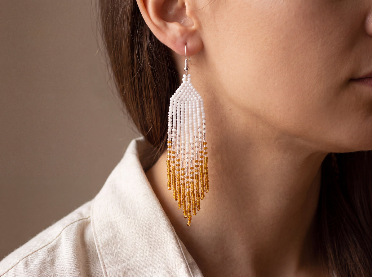 Handmade beaded fringe earring in white to amber-gold ombre worn on a model's ear, showing the long tassel drop.