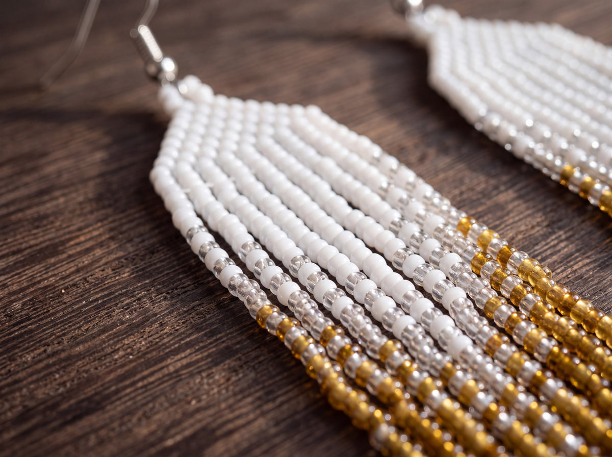Close-up of handmade white and gold beaded fringe earrings on dark wood, showing the ombre from white seed beads to amber tips.