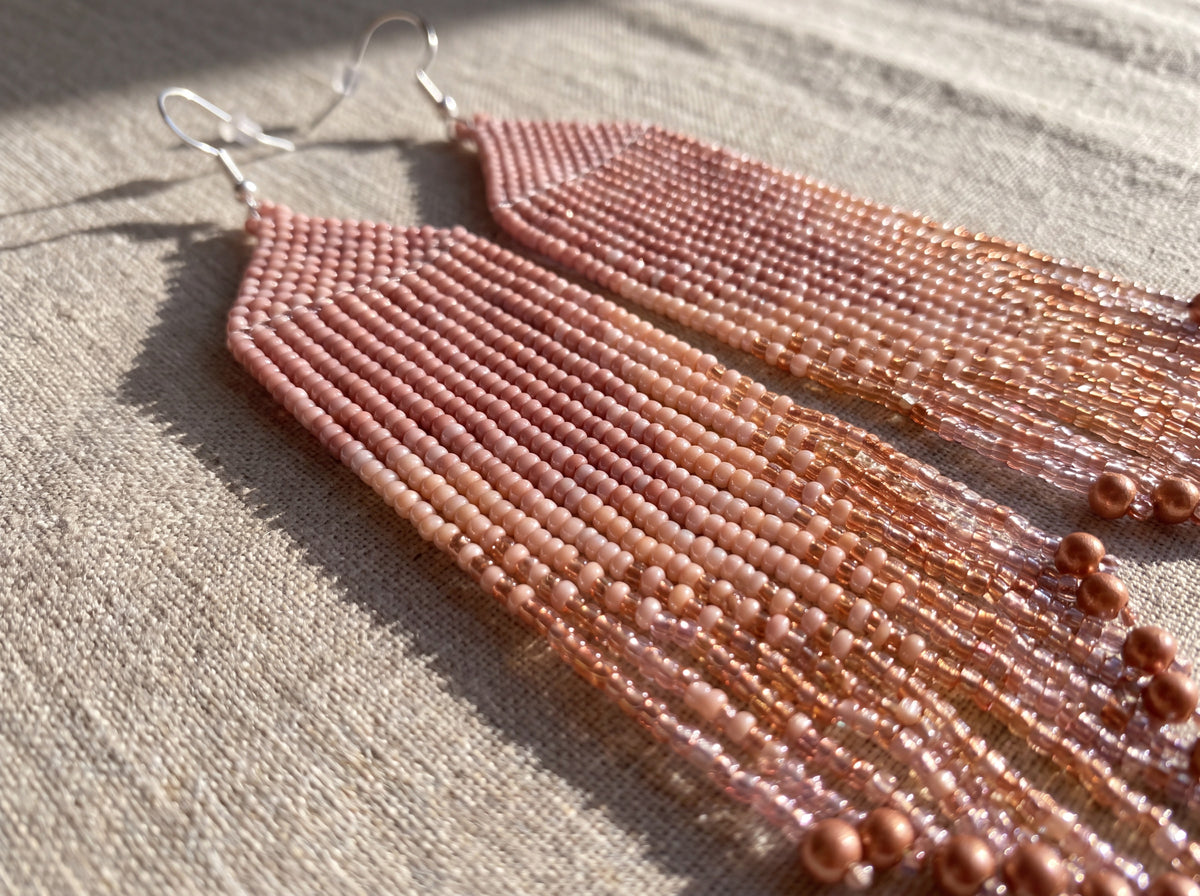 Close-up of long handmade beaded fringe earrings in blush pink and sparkling rose gold seed beads, laid flat on linen in sunlight