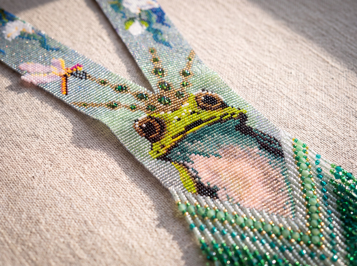 Close-up of a handmade beaded frog necklace showing the crowned frog portrait, dragonfly detail and emerald crystal drop fringe on linen.