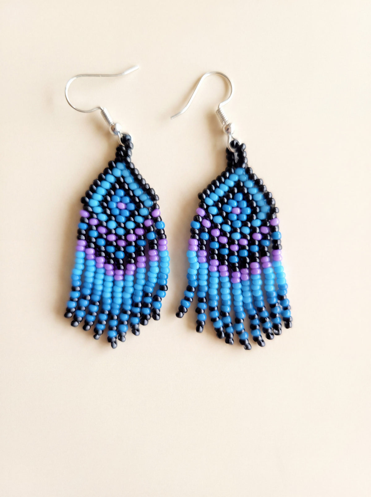 Handmade blue and black beaded fringe earrings with purple glass seed bead accents on a cream backdrop