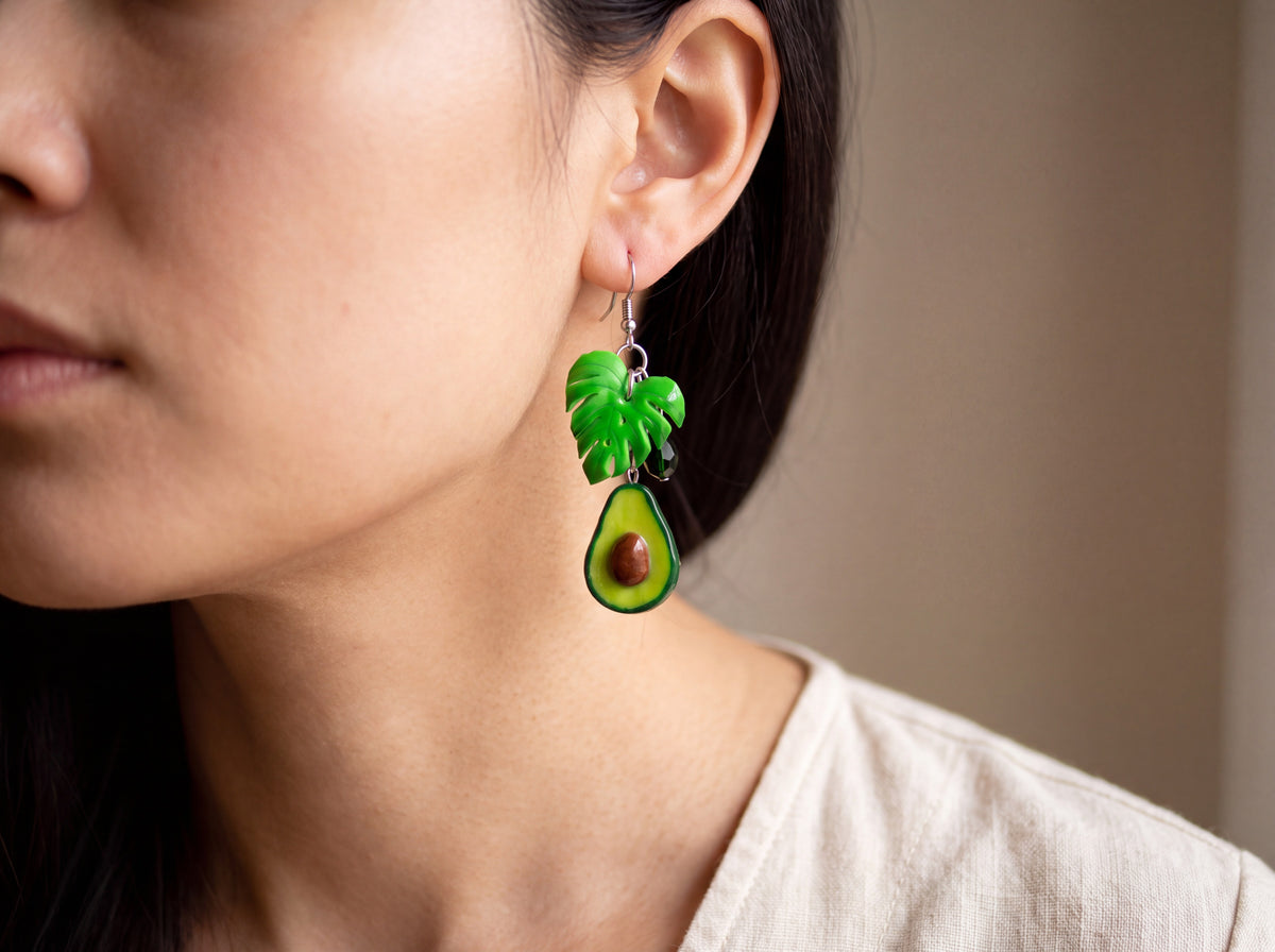 Handmade avocado half earring with a monstera leaf and green crystal on a silver hook, worn on a model's ear.