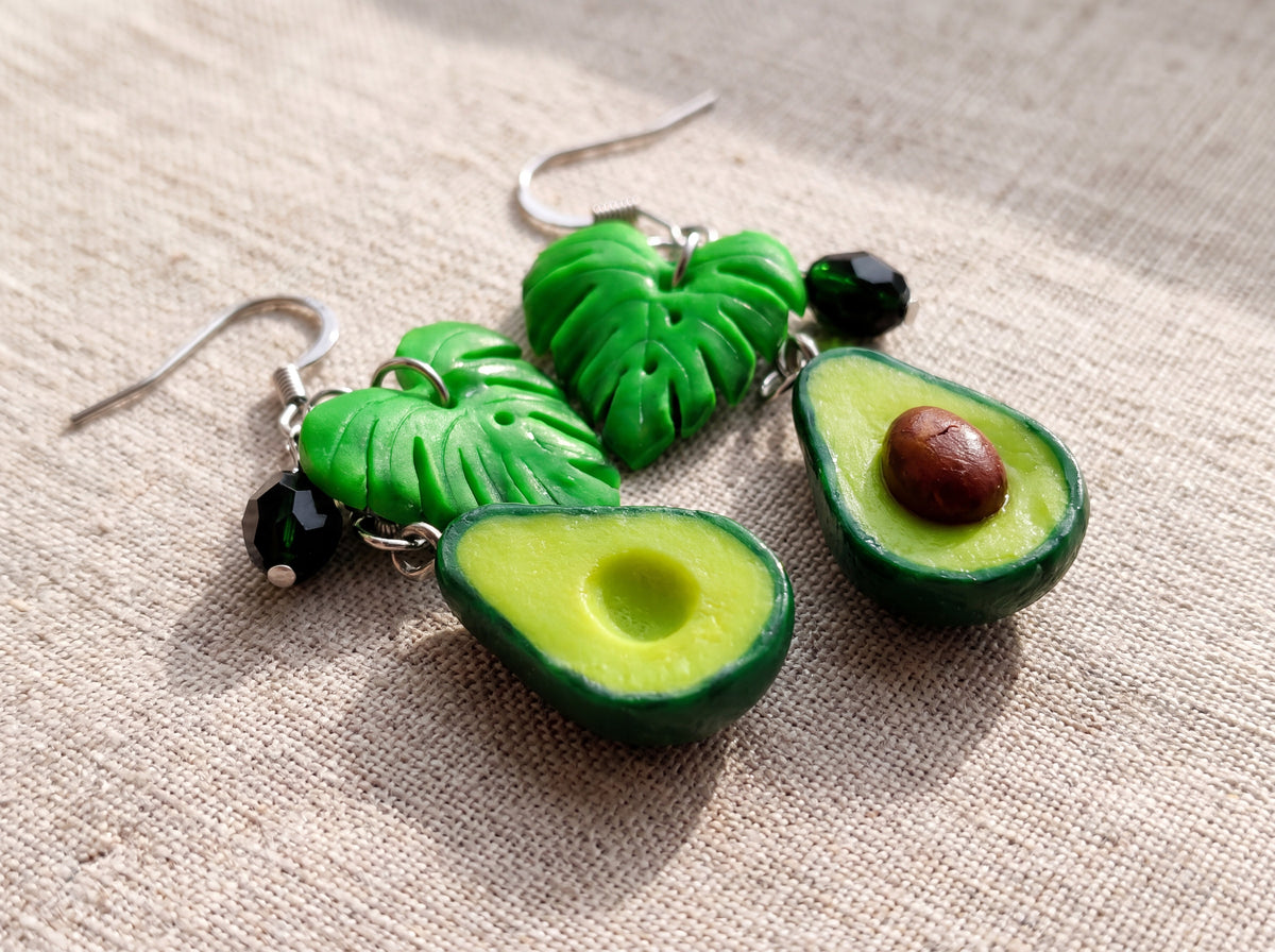 Close-up of handmade avocado half earrings with monstera leaves and dark green crystals on silver hooks against beige linen.