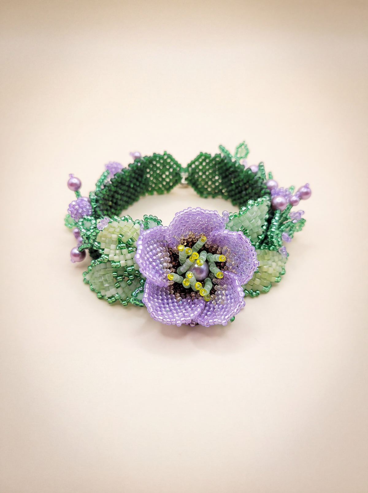 Handmade beaded bracelet with a purple flower centerpiece, green leaves and lavender pearl accents on a cream backdrop