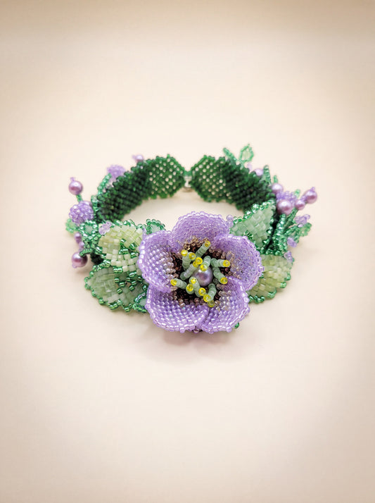 Handmade beaded bracelet with a purple flower centerpiece, green leaves and lavender pearl accents on a cream backdrop