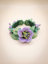 Handmade beaded bracelet with a purple flower centerpiece, green leaves and lavender pearl accents on a cream backdrop