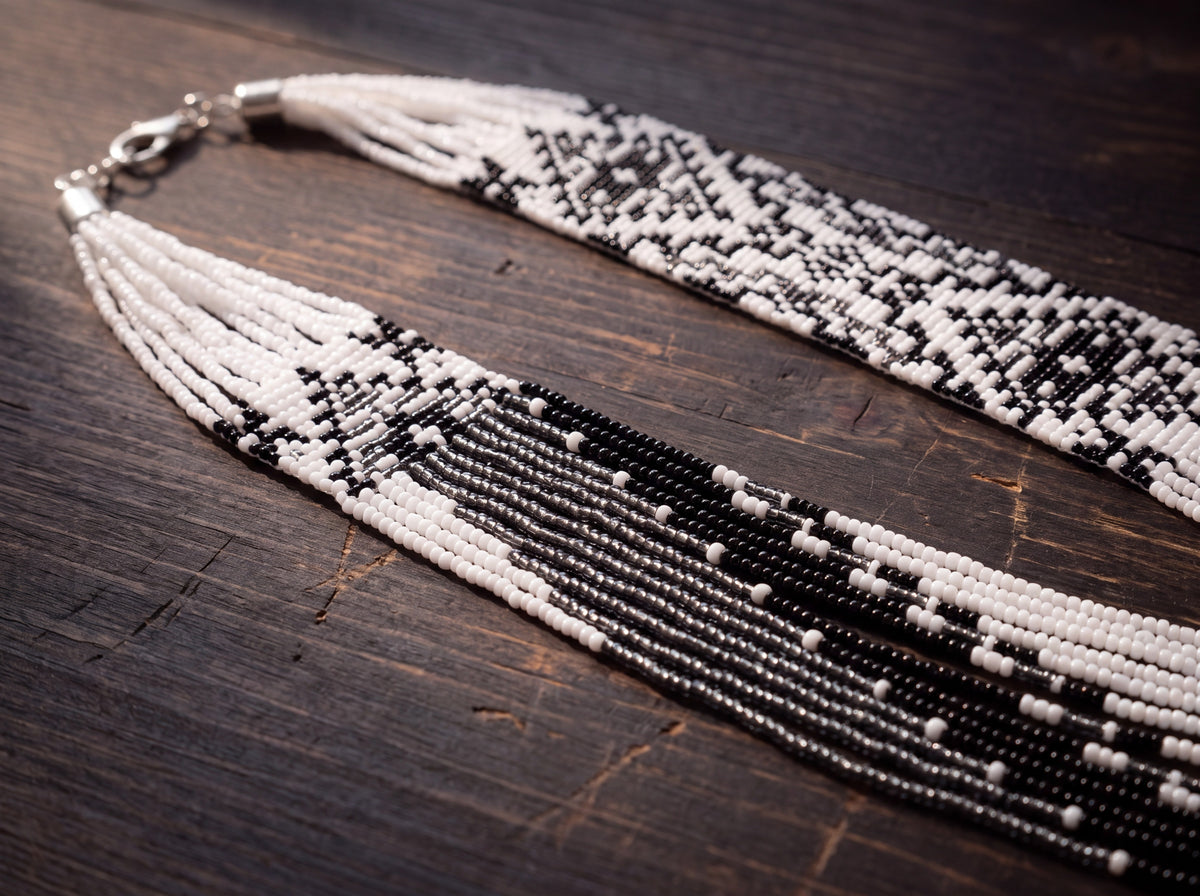 Close-up flat lay of a handmade beaded necklace with black, white, and silver seed bead strands and geometric woven panels on dark rustic wood.