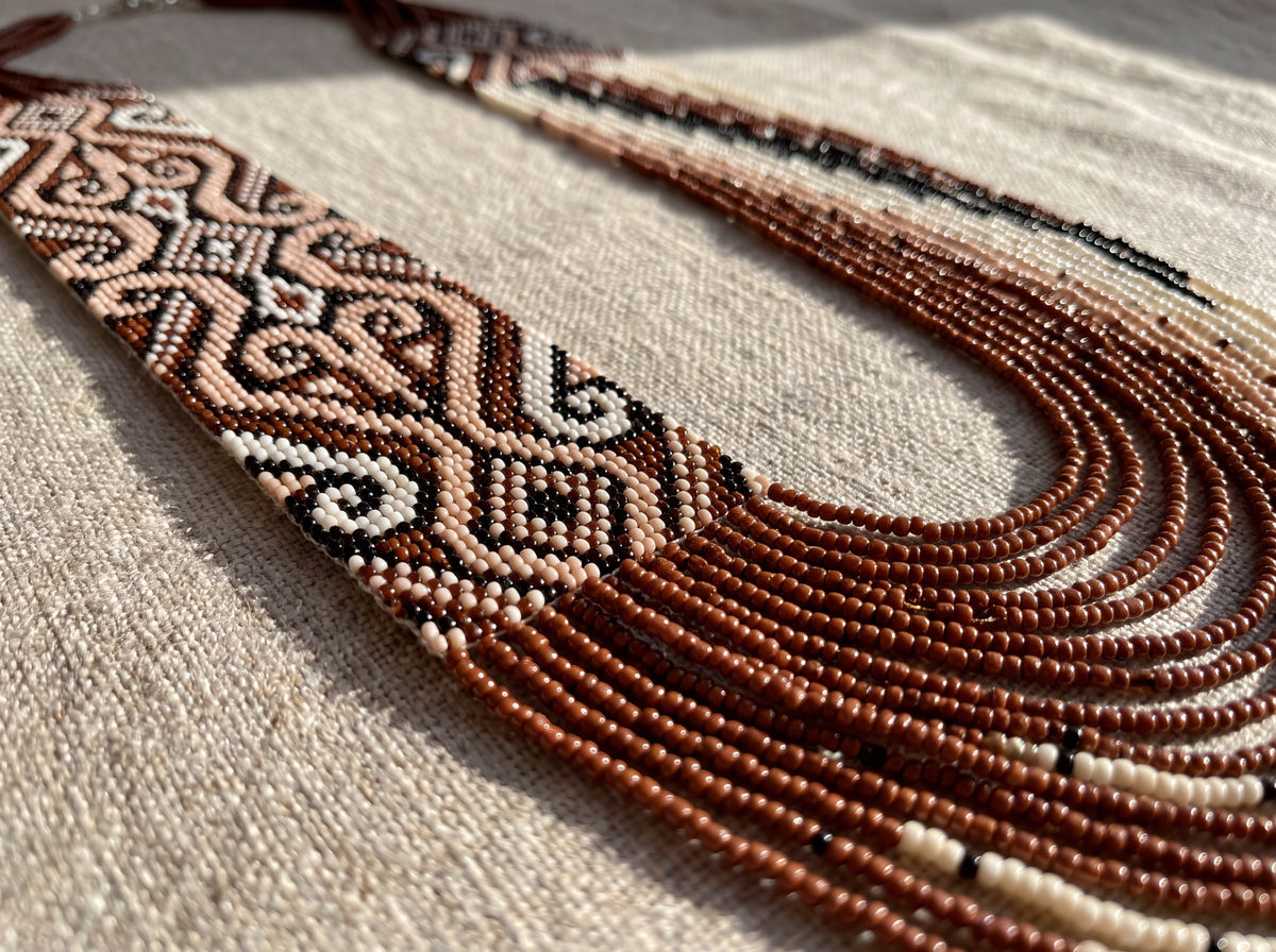 Close-up of a handmade beaded necklace showing a woven brown and blush ornamental panel and layered terracotta seed bead strands on linen fabric.
