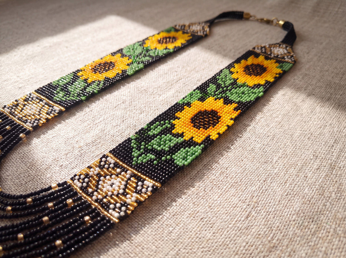 Close-up flat lay of a handmade beaded sunflower necklace showing woven yellow flowers, green leaves, and gold ornament bands on natural linen.