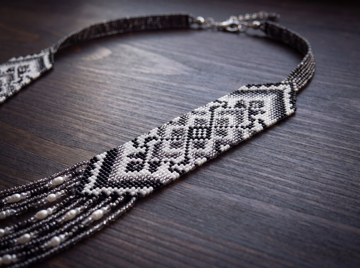 Close-up of a handmade black, white and silver beaded gerdan on dark wood, showing the woven ornament and multi-strand pearl fringe.