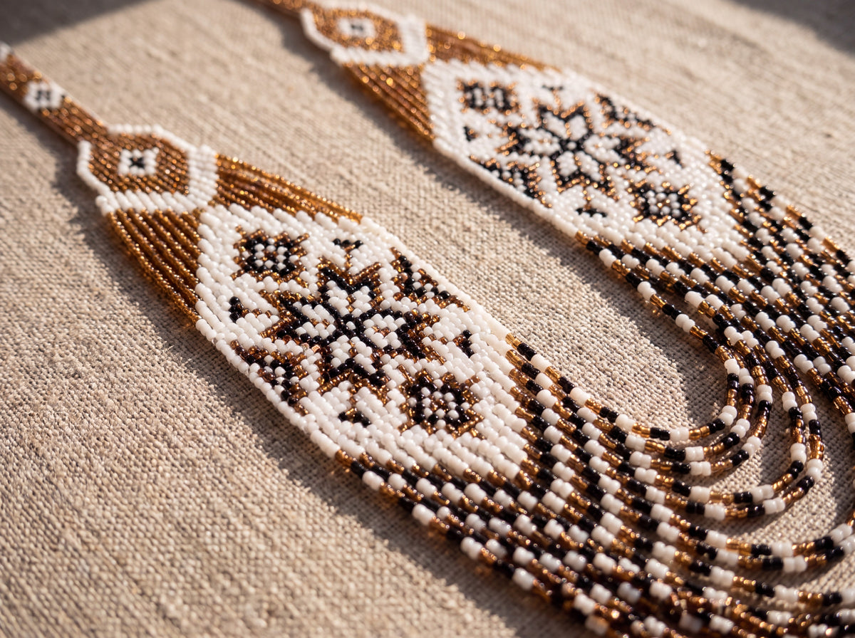 Detail of a handmade beaded gerdan with white and gold star pattern panels and draped bead strands, photographed on natural linen.