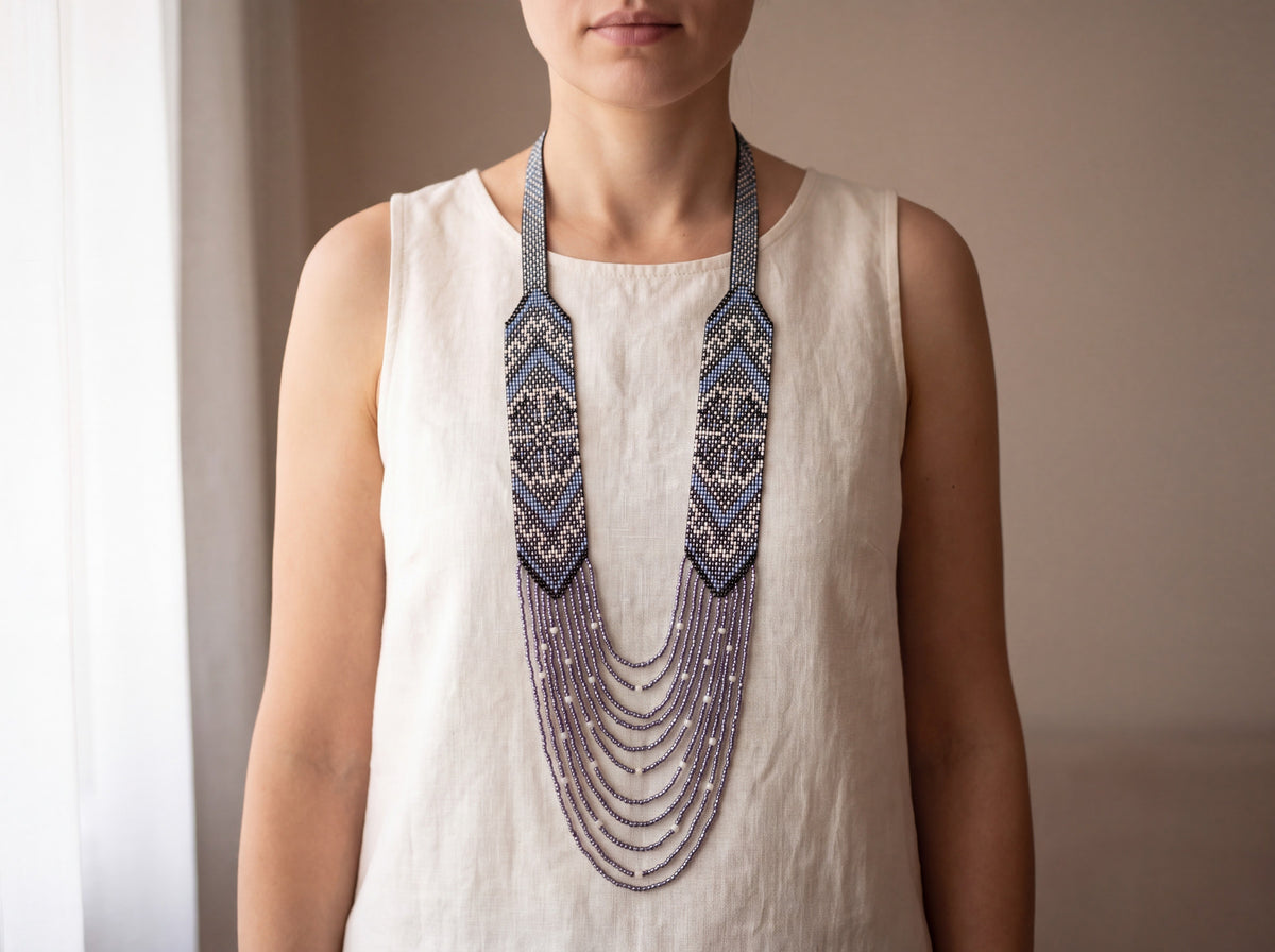 Handmade beaded gerdan necklace in blue, black and white geometric patterns with layered lavender strands, worn on a model in a cream linen top
